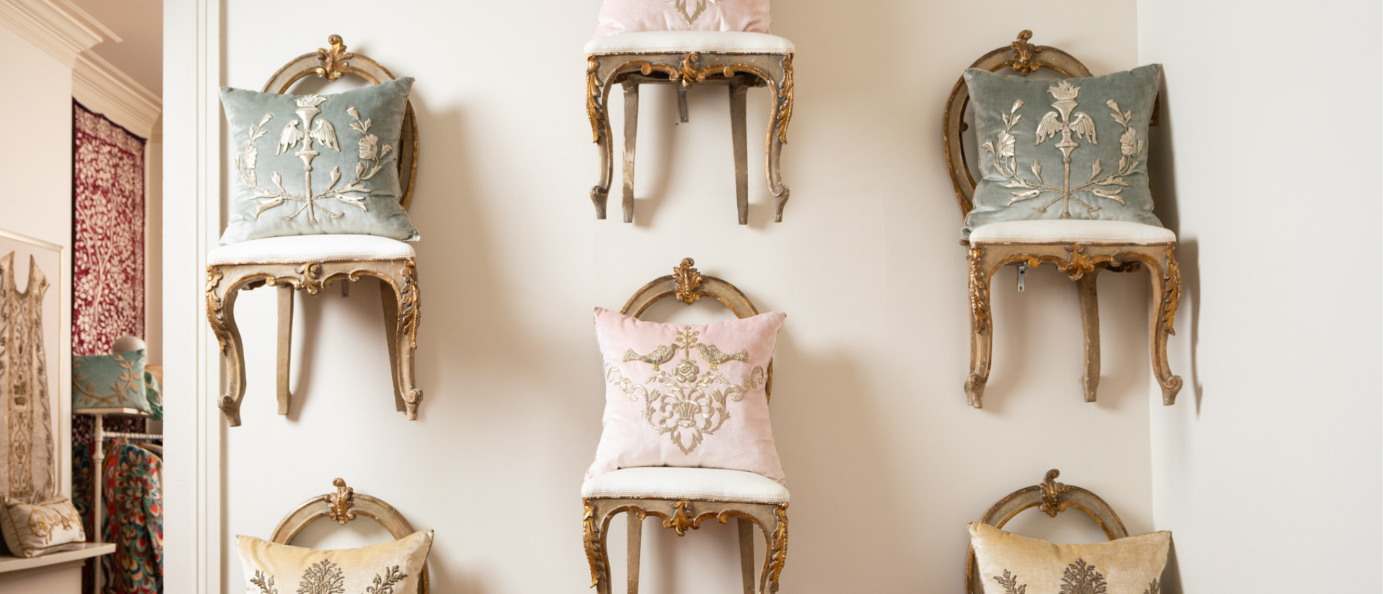 B. Viz Design New Orleans Atelier's famous Flying Chair Wall display features beautiful antique chairs on a white wall with extraordinary B. Viz Design antique textile square pillows in an array of colors and embroideries.